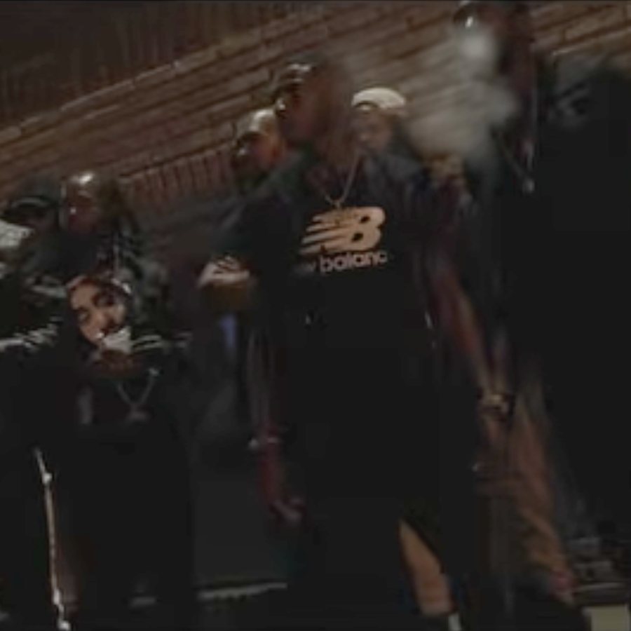 A still from the Bring the City Out video: Silent 777 in a black New Balance shirt, standing among a crowd in front of a brick wall at night.
