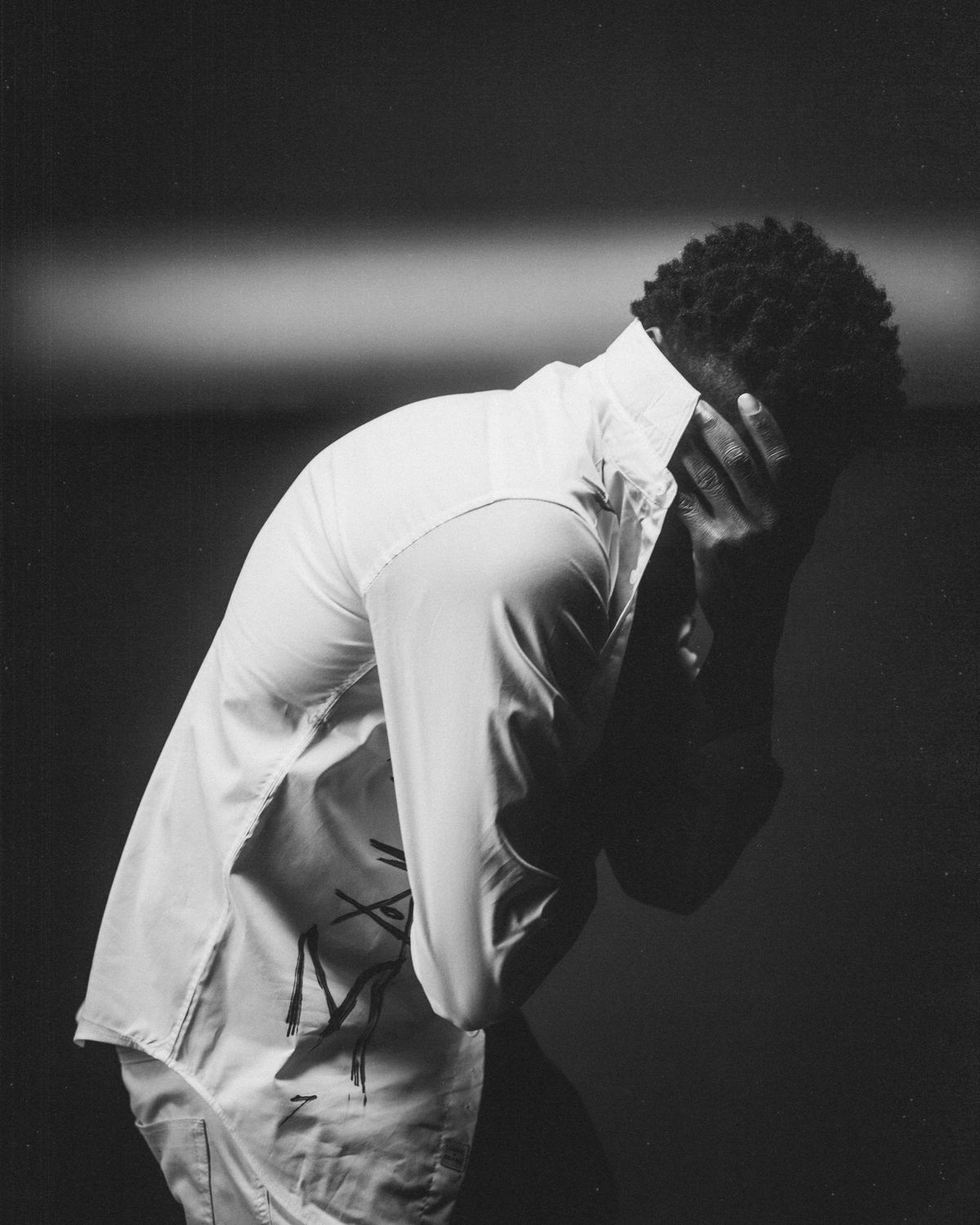 Black and white photo of Silent 777 bent forward in a white shirt with a hand covering his face.