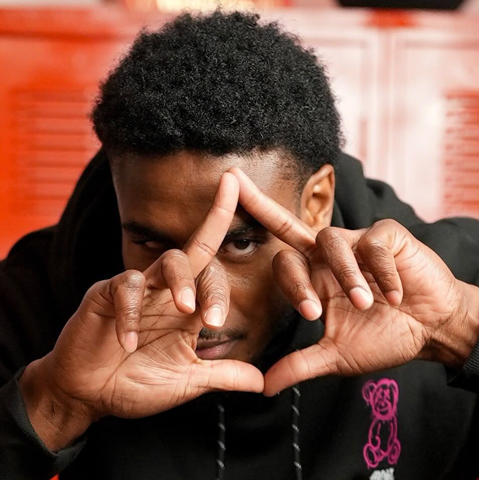 Silent 777 making a triangle with his hands in front of one eye, red lockers behind him.