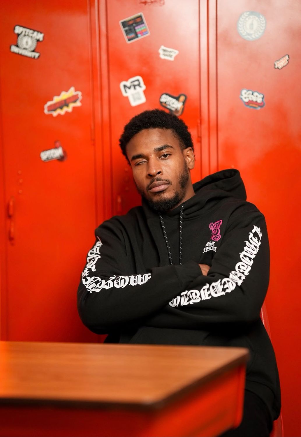 Silent 777 seated at a school desk in front of red lockers covered in stickers, arms crossed, in a black hoodie with white lettering on the sleeves.