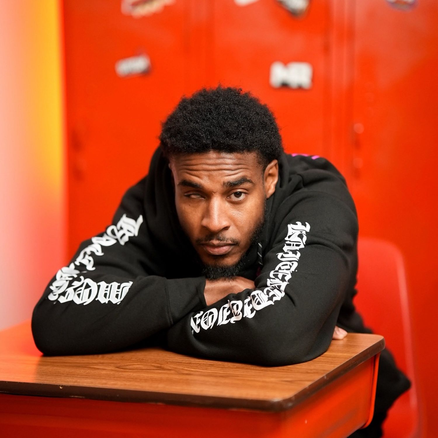 Silent 777 leaning on a desk with his arms folded in front of red lockers.