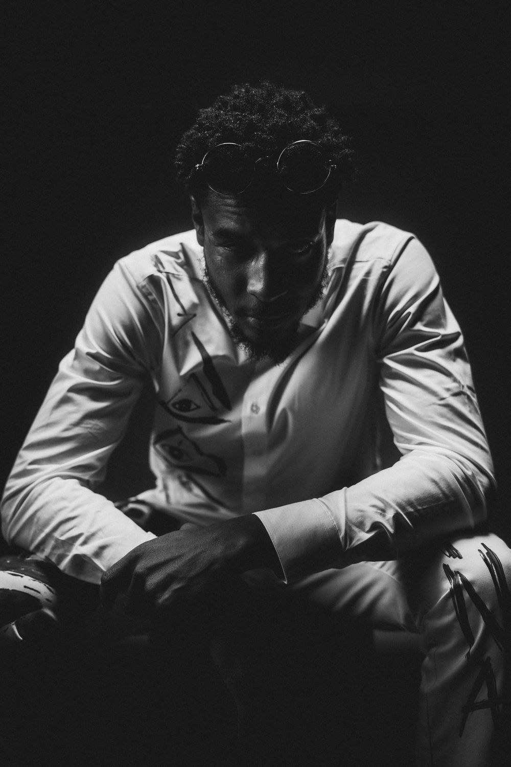 Black and white photo of Silent 777 seated in dim light, round glasses resting on his head, in a white shirt with drawn eye designs.
